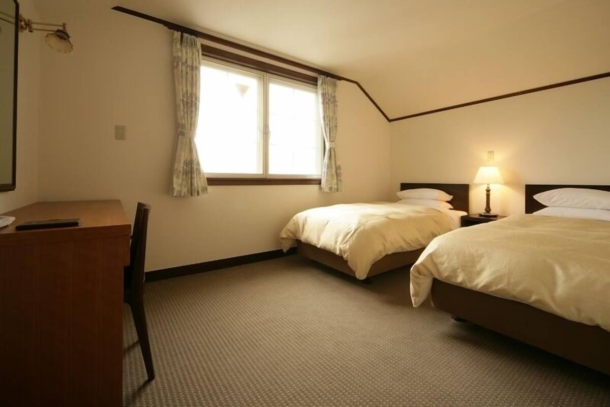 A hotel room with two beds, a desk, a lamp, and a window with curtains.