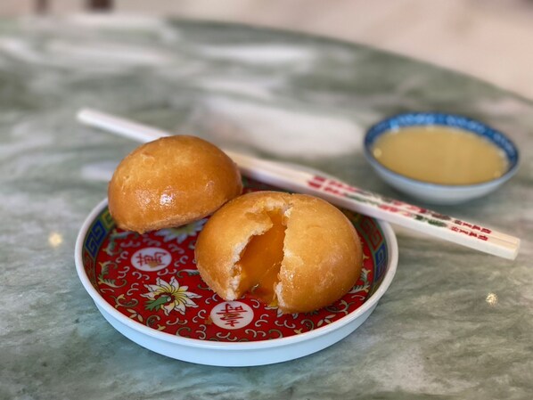 Une assiette avec deux pâtisseries rondes, dorées et légèrement brunes, et un petit bol de sauce jaune, le tout accompagné de baguettes.