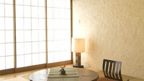 Une pièce avec un sol recouvert de tatamis, comprenant une table ronde, deux chaises, une lampe et une vue sur l’extérieur à travers des panneaux coulissants.