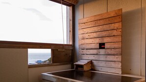 Uma onsen japonesa tradicional com um banco de madeira, uma pequena construção de madeira e vista para o mar através de uma janela.