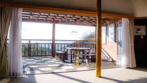 Quarto com teto de madeira, uma janela grande com cortinas, vista para o exterior e uma mesa de madeira com cadeiras.