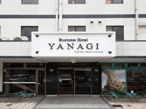 Eksteriøret af et forretningshotel kaldet YANAGI, en del af Yutaka Hotel Group. Det har en iøjnefaldende skiltning, en glasindgang og en parkeringsplads med biler.