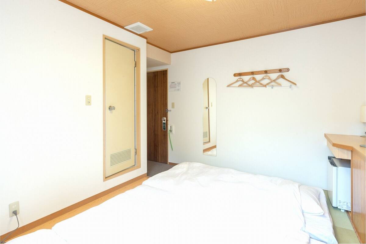 Habitación de hotel con una cama, cabecero de madera, espejo, perchero y un pequeño escritorio con lámpara.