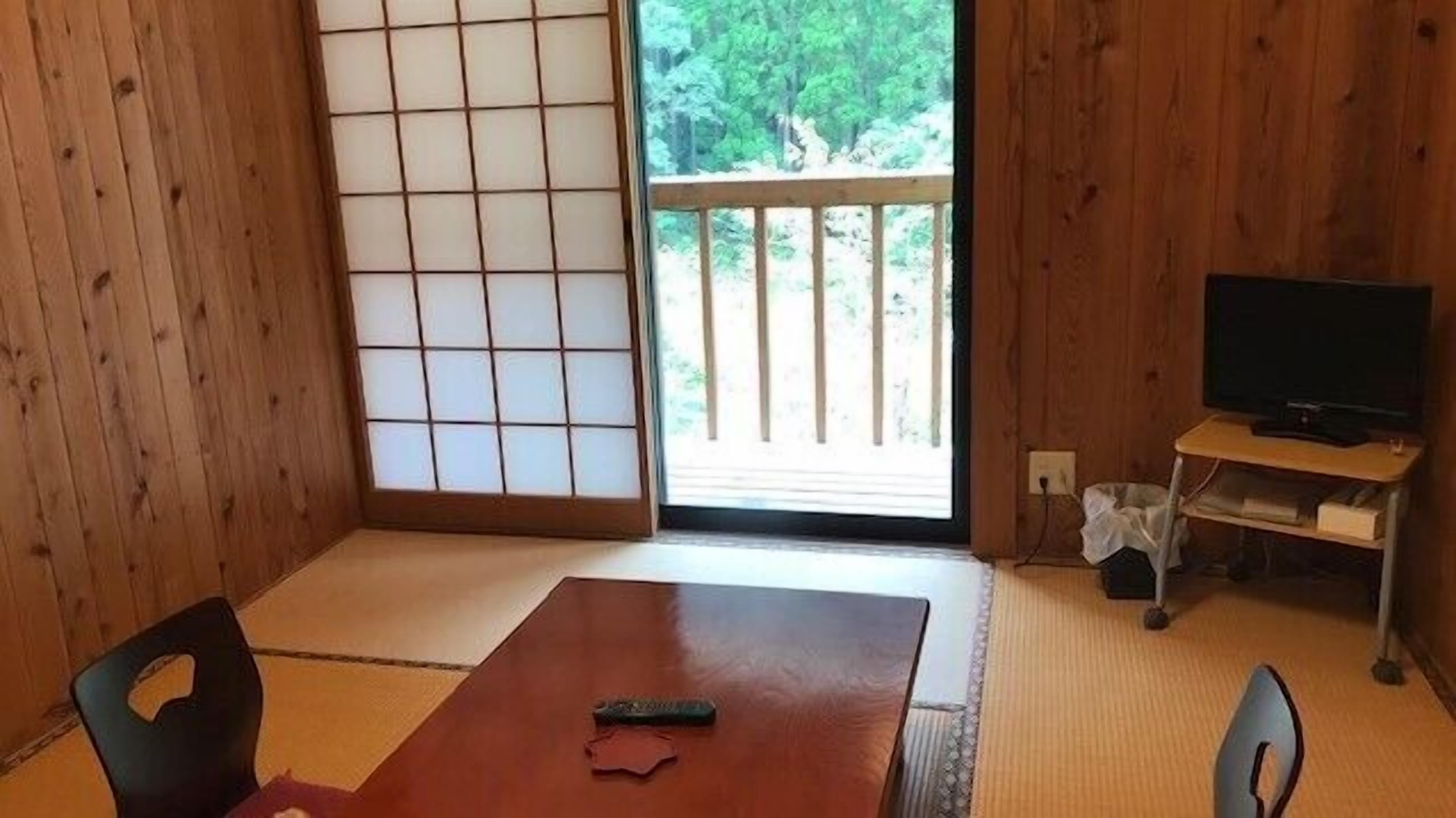 A room with tatami flooring, a sliding shoji screen, a TV on a stand, and a view of a balcony with trees.