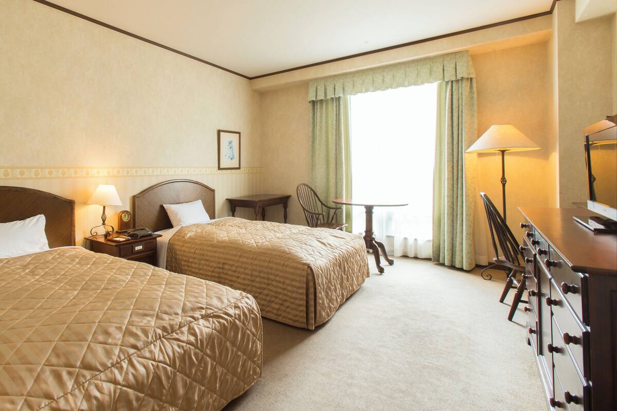Una habitación de hotel con dos camas, un escritorio, una silla, un televisor y una ventana con cortinas.