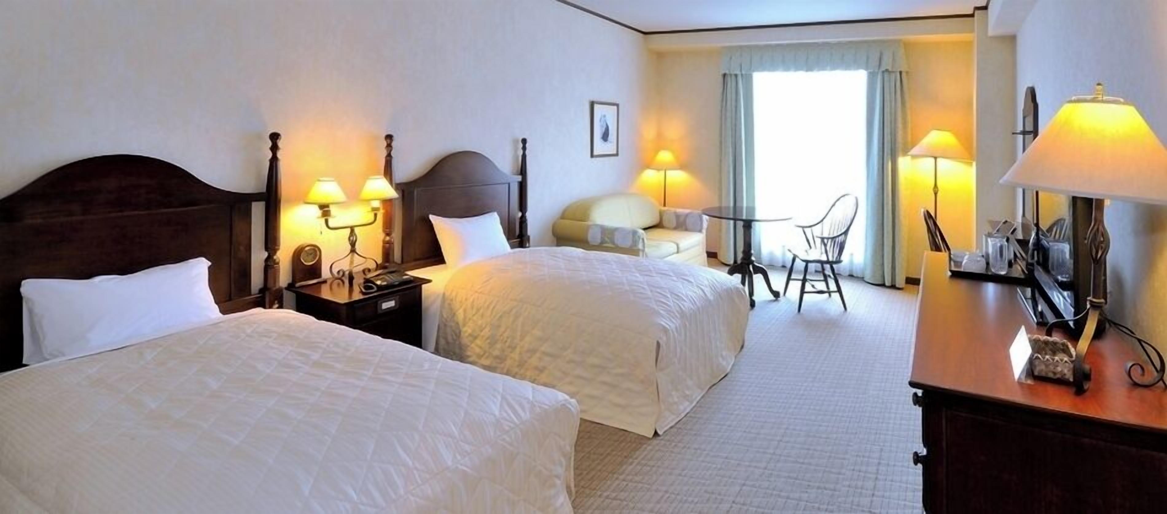 Quarto de hotel com duas camas, mesas de cabeceira com abajures, uma mesinha com cadeiras e vista para o exterior através de uma janela.