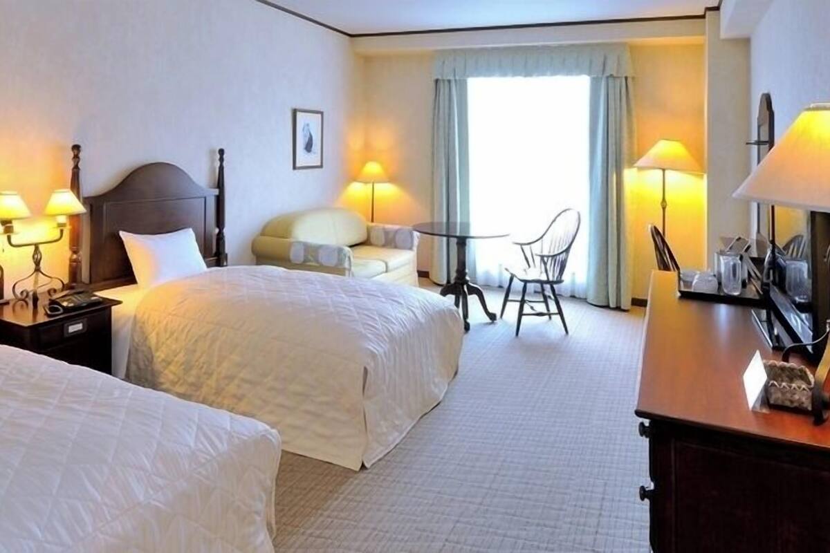Una habitación de hotel con dos camas, mesitas de noche con lámparas, una pequeña mesa con sillas y vistas al exterior a través de una ventana.