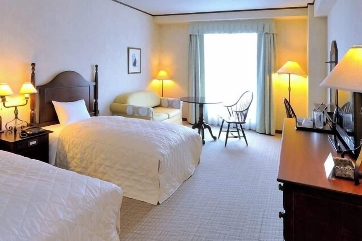 Ein Hotelzimmer mit zwei Betten, Nachttischen mit Lampen, einem kleinen Tisch mit Stühlen und Blick durch ein Fenster nach draußen.