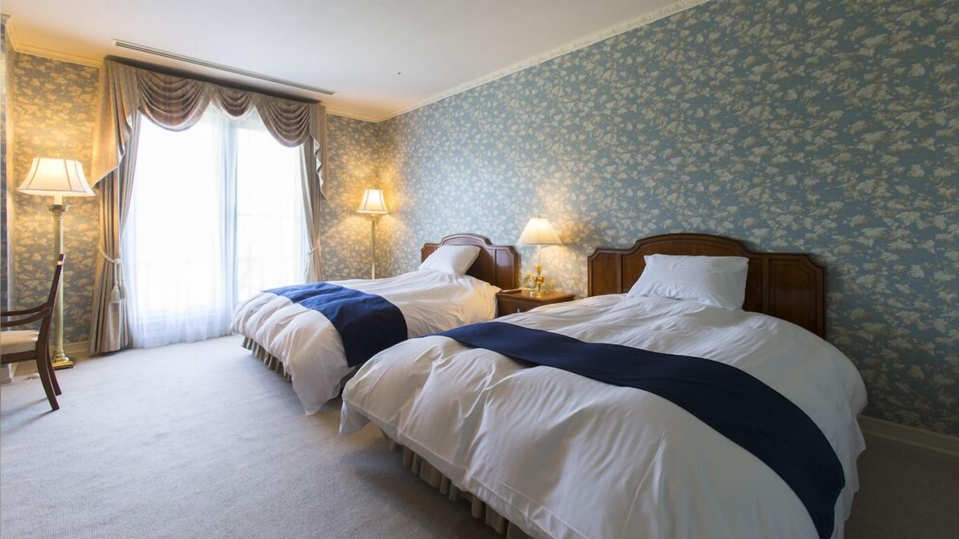 Quarto de hotel com duas camas, uma janela grande com cortinas, um abajur e papel de parede floral.