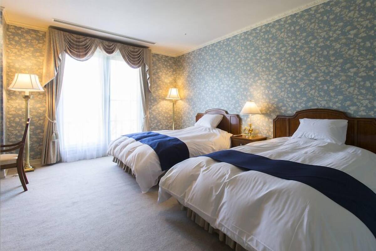 Habitación de hotel con dos camas, un ventanal con cortinas, una lámpara y un papel tapiz floral.