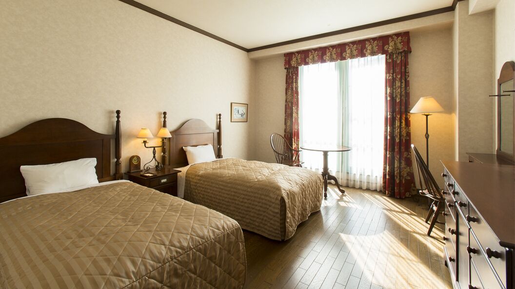 Quarto de hotel com duas camas, uma escrivaninha com luminária, uma cadeira e uma janela com cortinas.