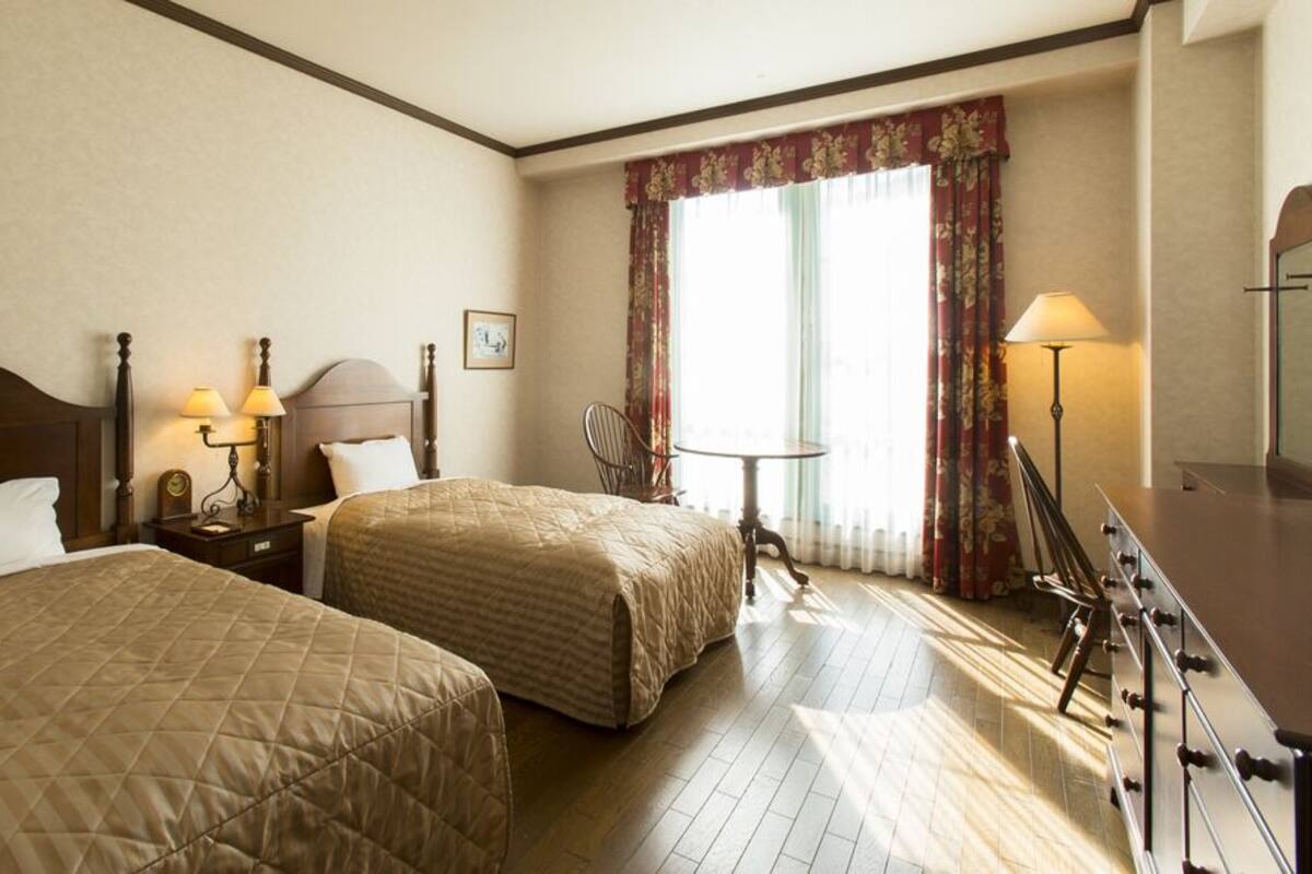 Habitación de hotel con dos camas, un escritorio con lámpara, una silla y una ventana con cortinas.