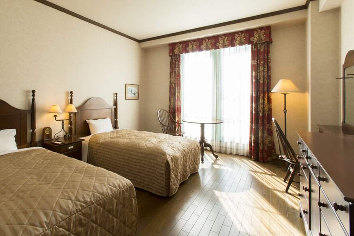Ein Hotelzimmer mit zwei Betten, einem Schreibtisch mit Lampe, einem Stuhl und einem Vorhangfenster.