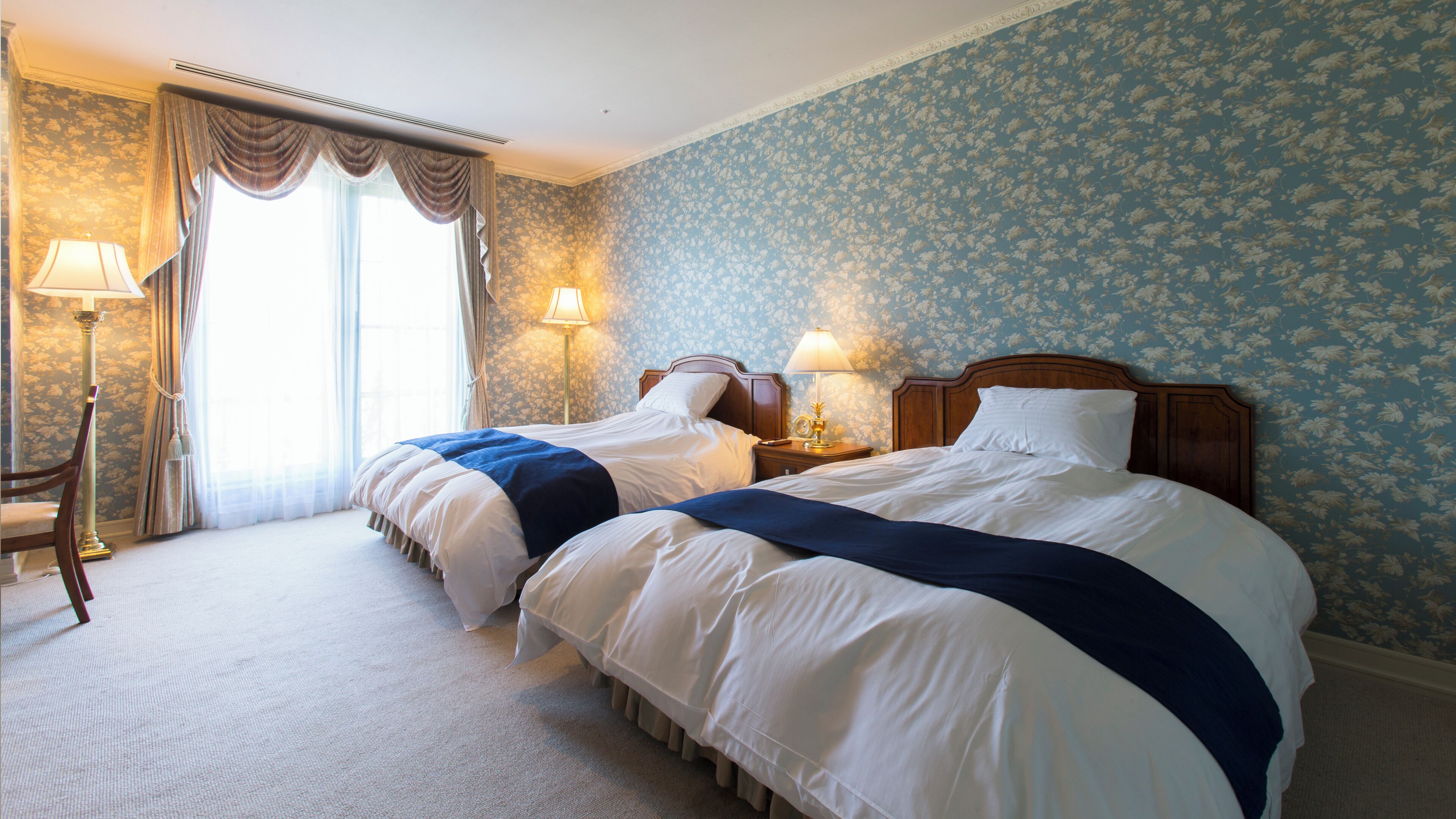 Quarto de hotel com duas camas, um espaço de estar com uma poltrona, luminária e janela com cortinas.
