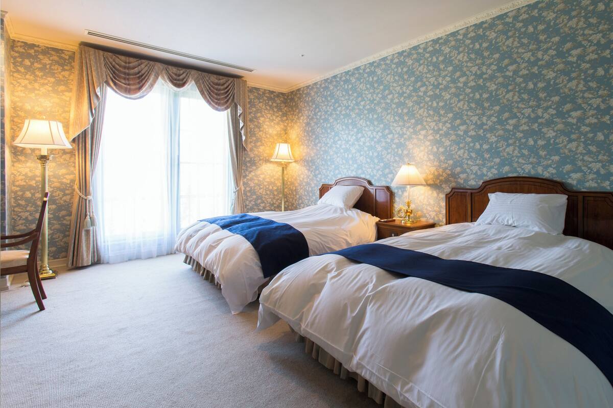 Habitación de hotel con dos camas, una zona de estar con una silla, una lámpara y un ventanal con cortinas.