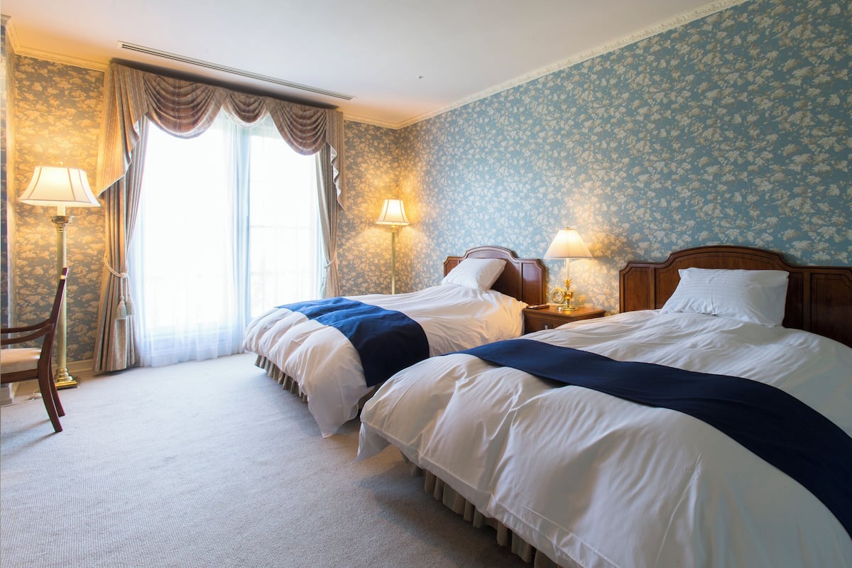 Ein Hotelzimmer mit zwei Betten, einer Sitzecke inklusive Sessel, einer Lampe und einem Fenster mit Vorhängen.
