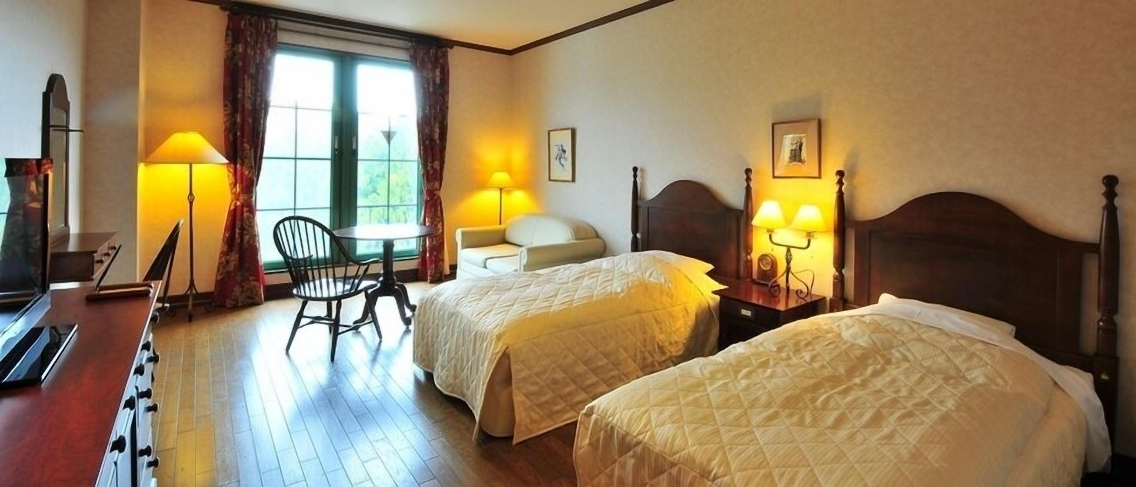 Quarto de hotel com duas camas, uma escrivaninha, uma cadeira e uma janela com cortinas.