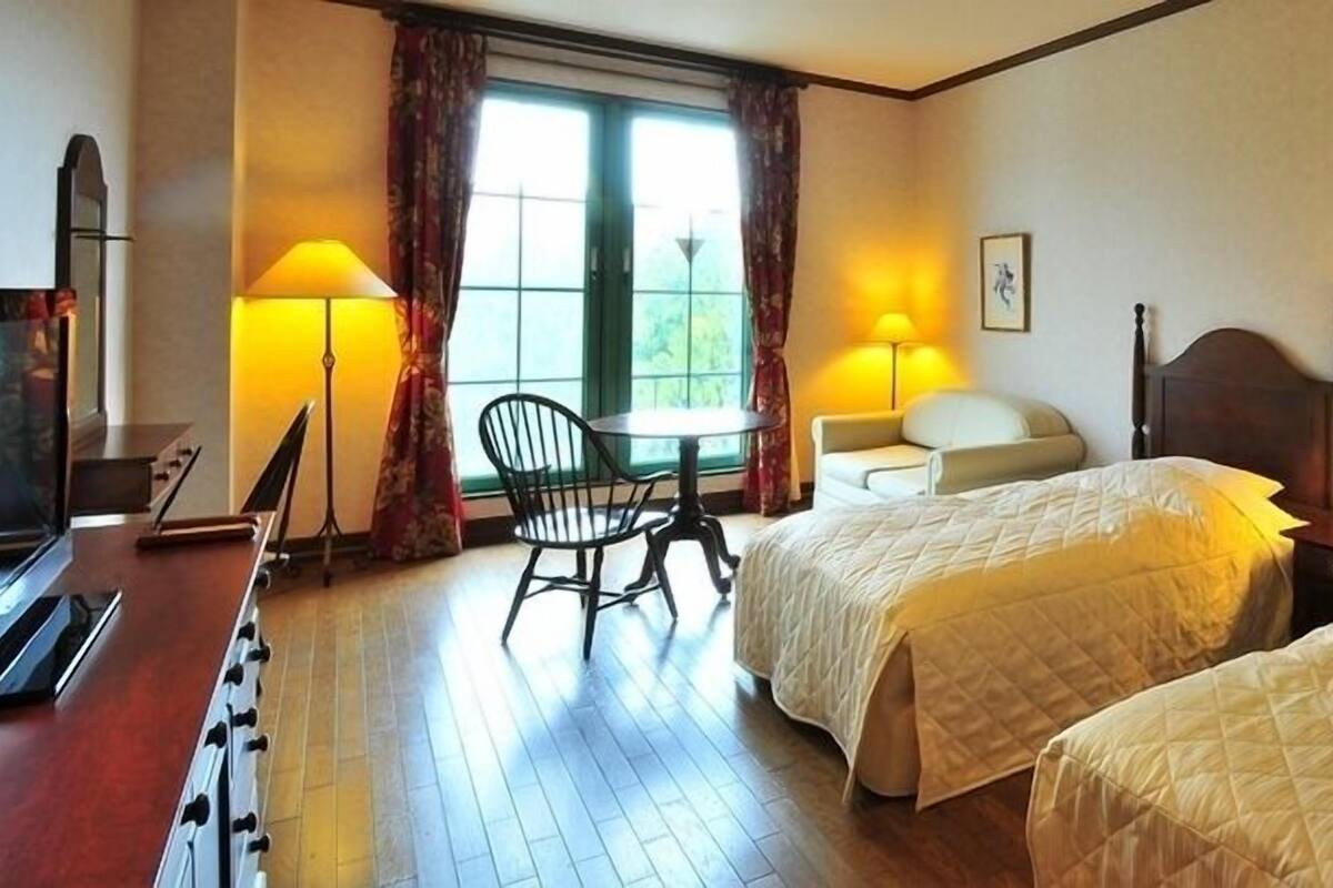 Habitación de hotel con dos camas, un escritorio, una silla y un ventanal con cortinas.