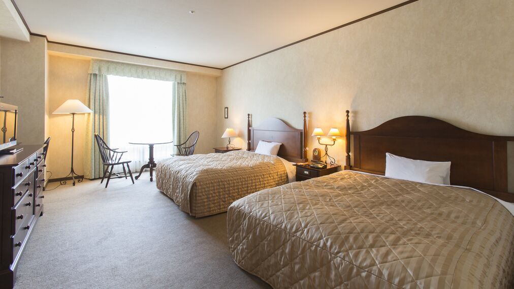 Quarto de hotel com duas camas, uma escrivaninha, uma cadeira e um abajur.