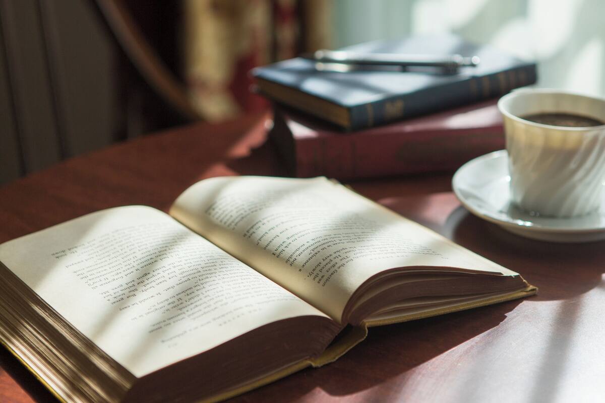 Un libro abierto sobre una mesa de madera, con una taza de café y otro libro al fondo.