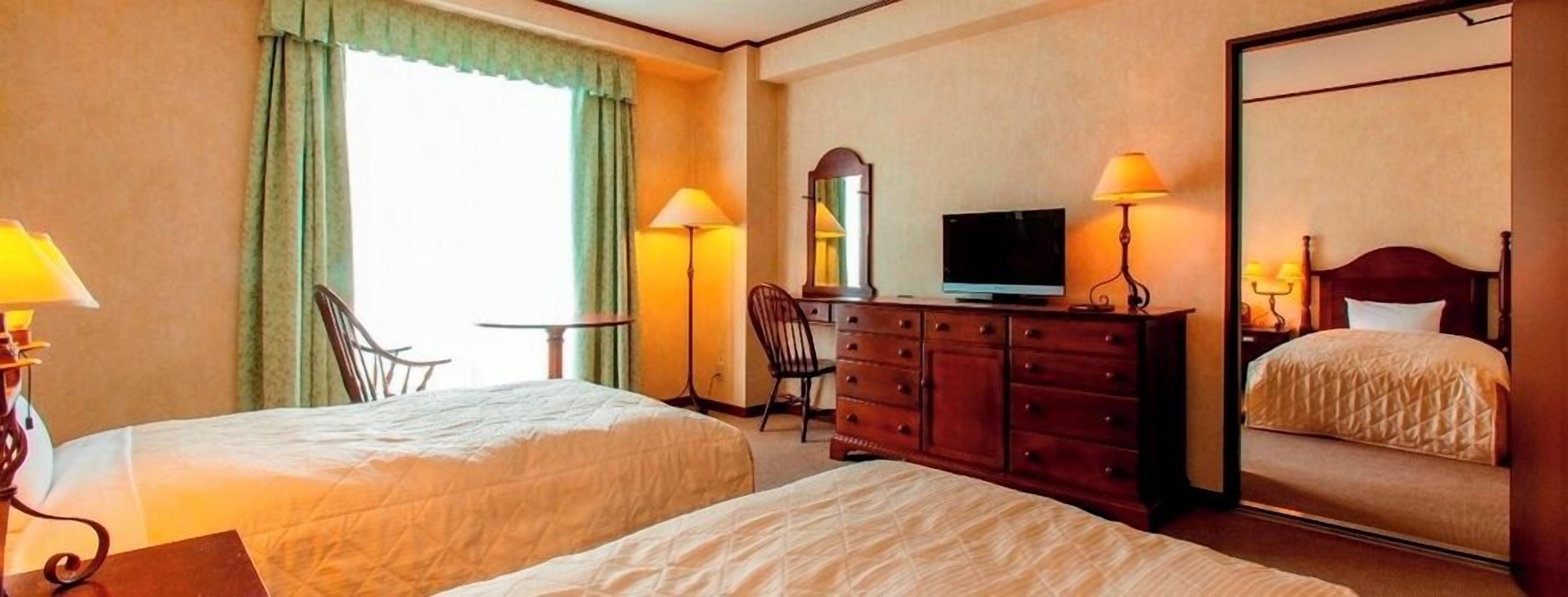 Ein Hotelzimmer mit zwei Betten, einem Fernseher, einem Schminktisch, einem Sessel und einem Spiegel.