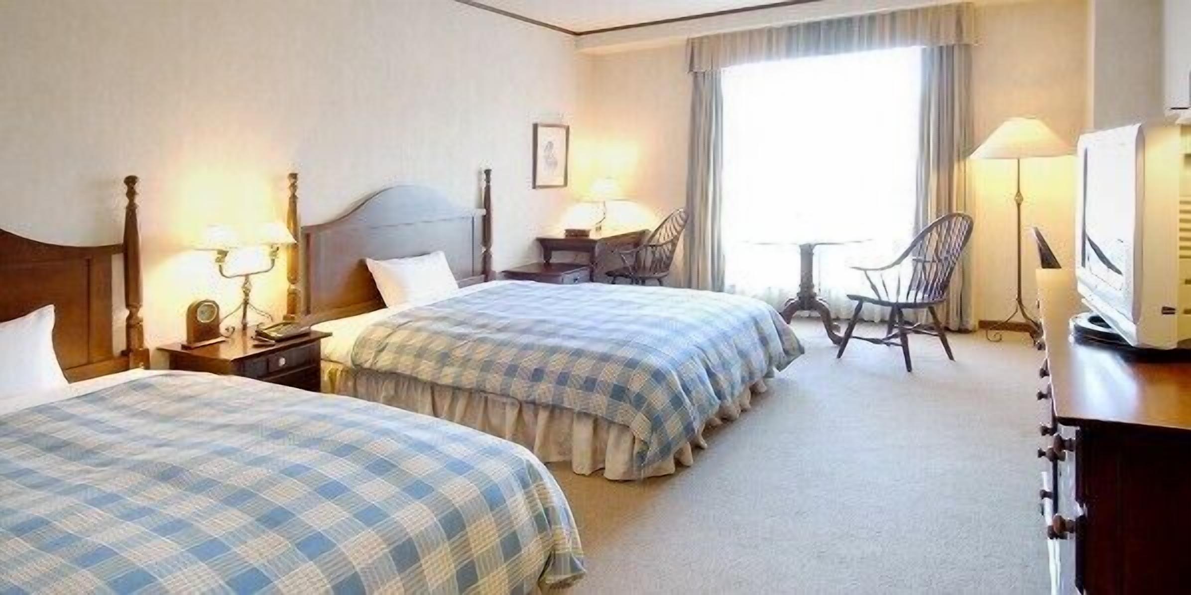 Quarto de hotel com duas camas, uma escrivaninha, uma cadeira, um abajur e uma televisão.
