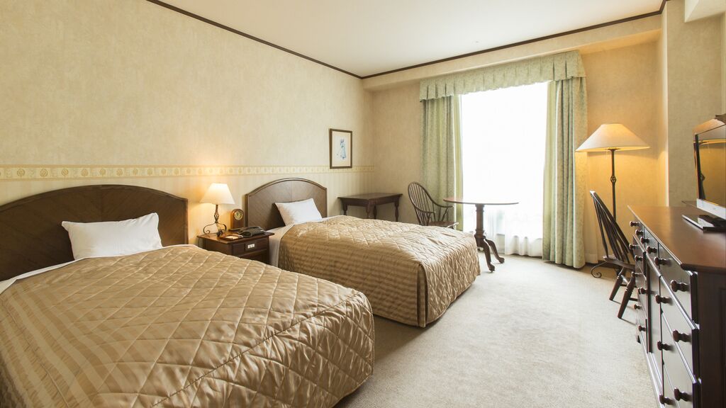Quarto de hotel com duas camas, uma escrivaninha, uma cadeira, um abajur e um aparador.