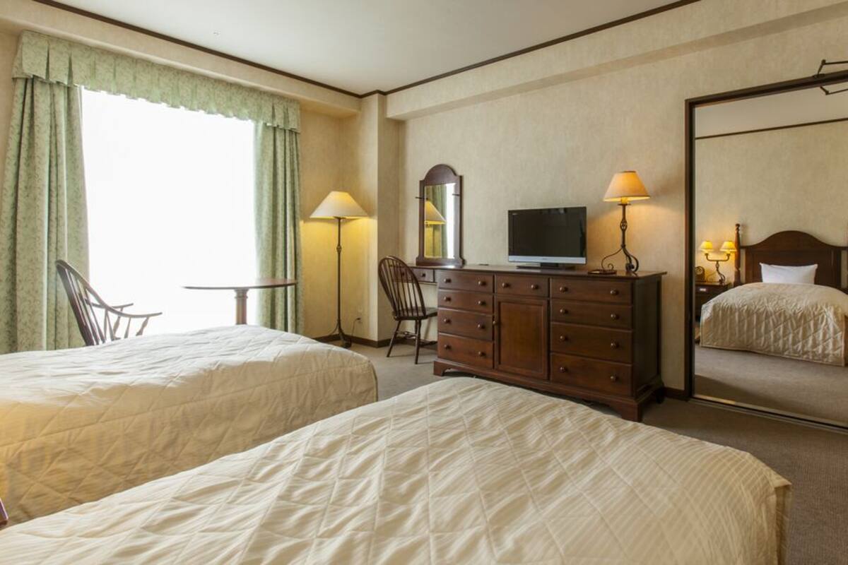 Habitación de hotel con dos camas, televisor, cómoda, espejo y ventana con cortinas.