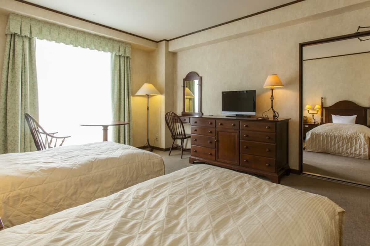 Ein Hotelzimmer mit zwei Betten, einem Fernseher, einem Schminktisch, einem Spiegel und einem Fenster mit Vorhängen.