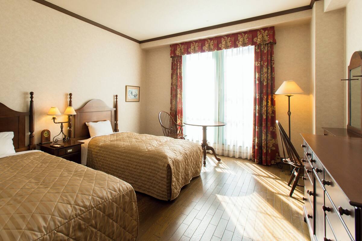 Habitación de hotel con dos camas, una mesita de noche con lámparas, una silla, una pequeña mesa y un aparador.