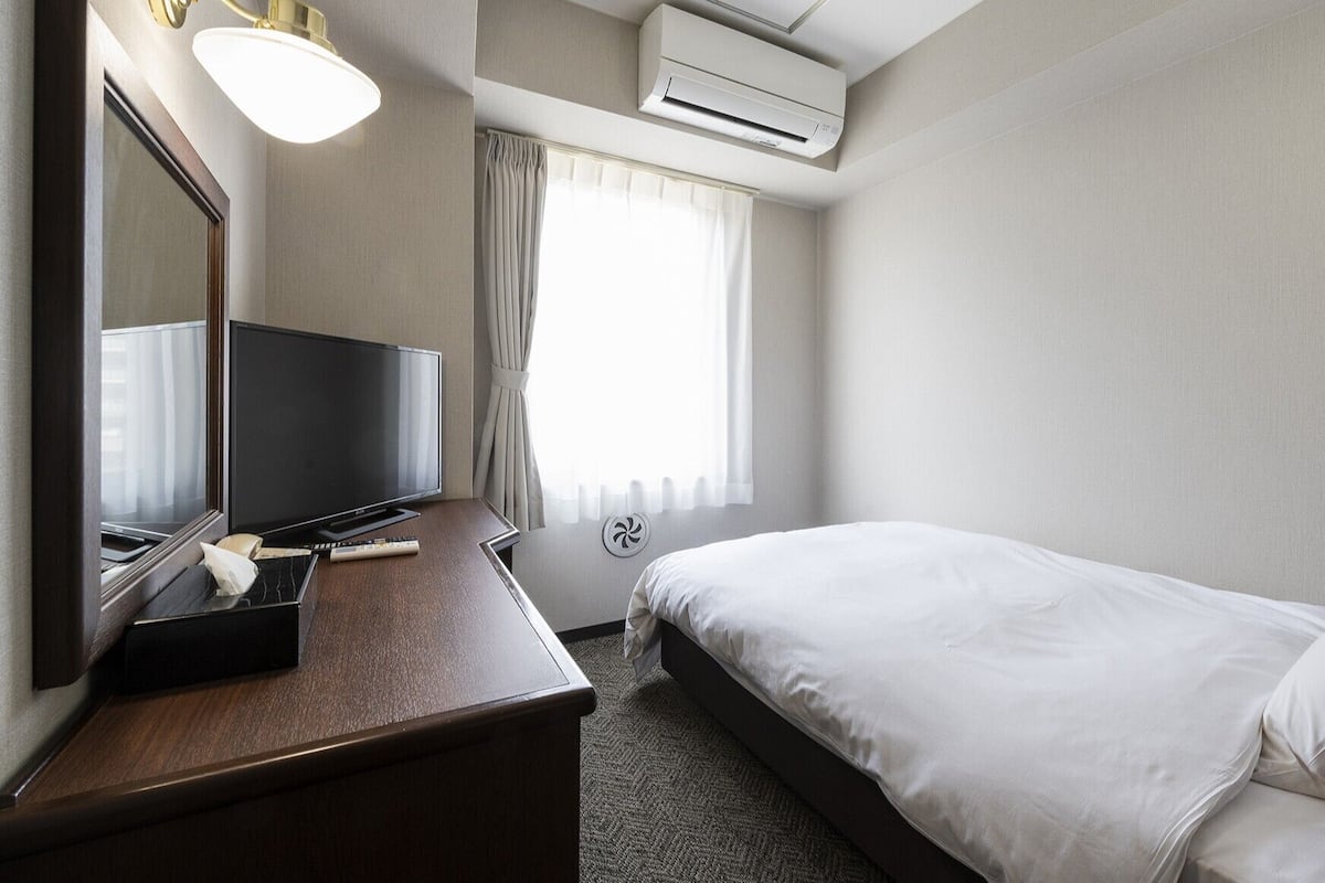 A hotel room with a bed, a TV, a desk, a mirror, an air conditioner, and a window with curtains.