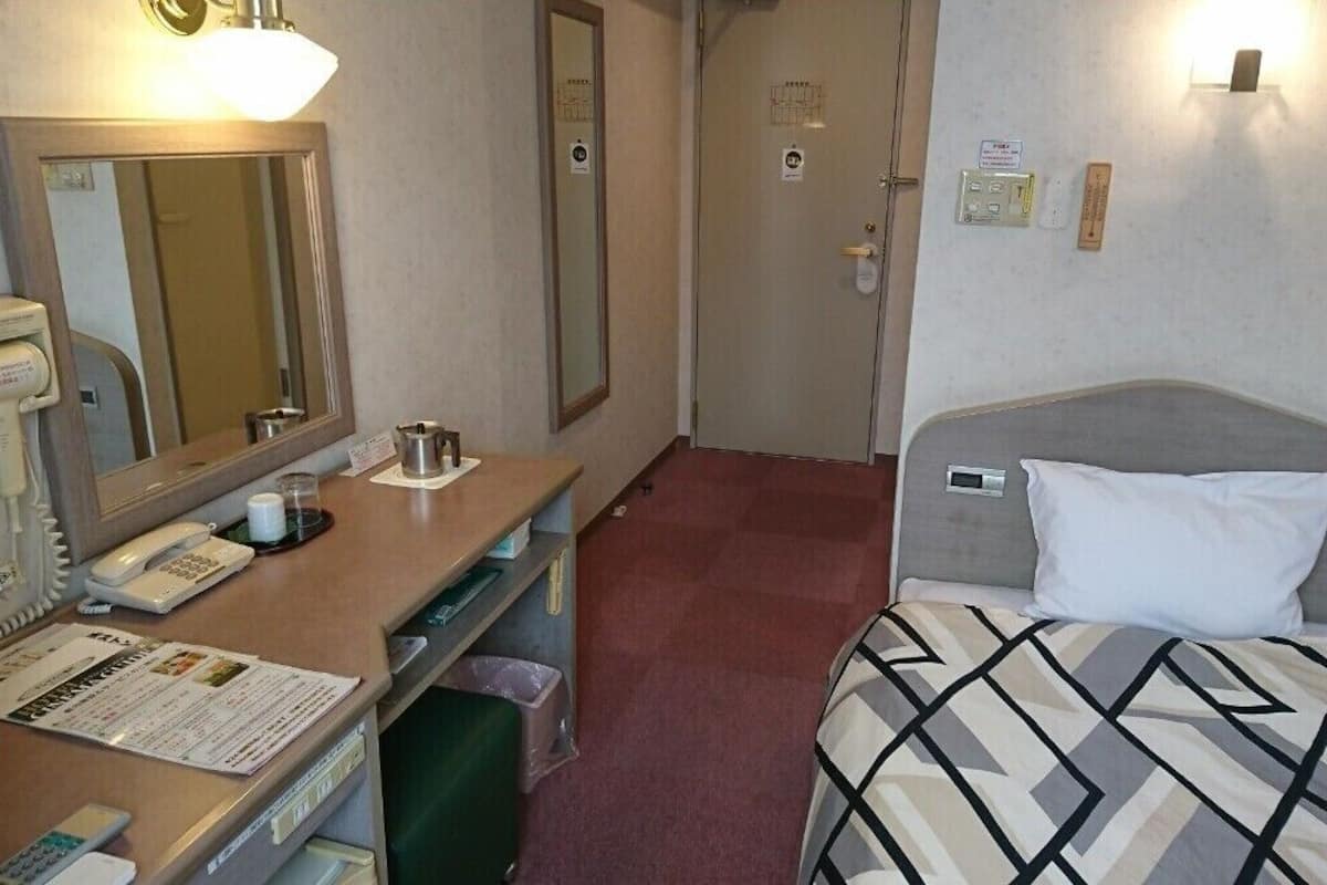 A hotel room with a bed, a desk with a telephone, a mirror, and a wall-mounted light fixture.