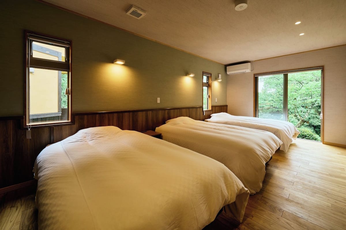 A room with two beds, wooden flooring, and large windows offering a view of greenery.