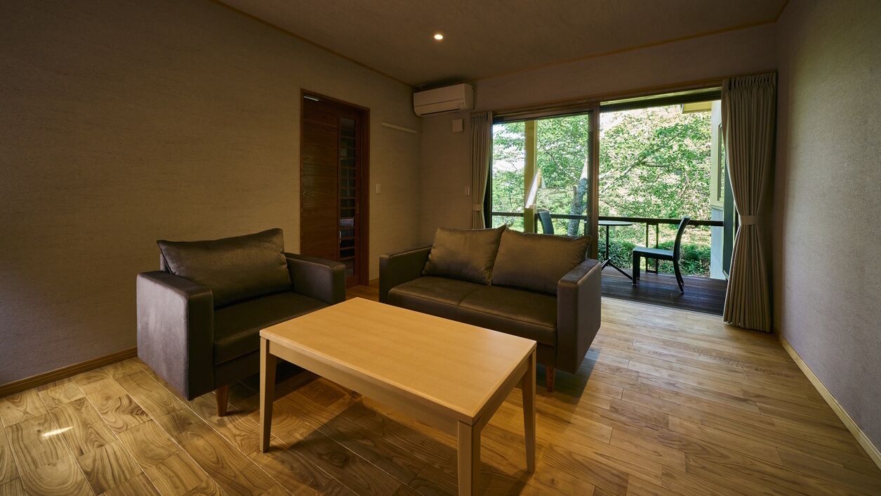 Habitació amb sòl de fusta, un sofà, una cadira, una taula i una finestra amb vista als arbres.