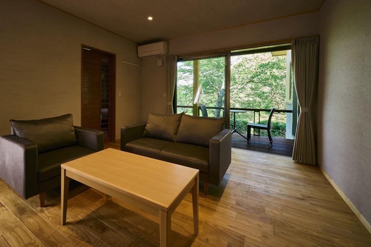 Habitació amb sòl de fusta, un sofà, una cadira, una taula i una finestra amb vista als arbres.