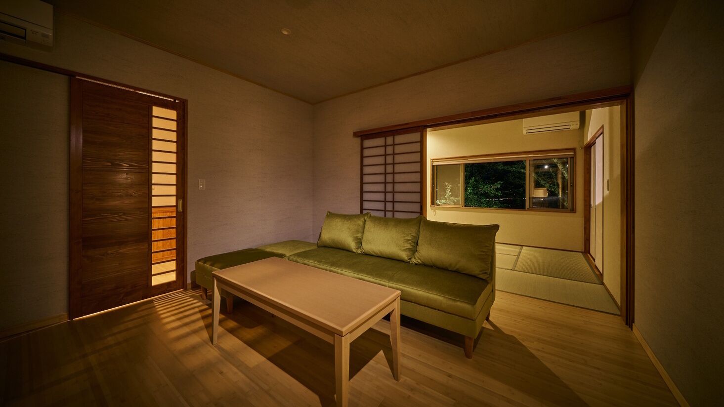 Quarto com piso de madeira, sofá verde, mesa de madeira, porta deslizante e janela com vista para área verde.