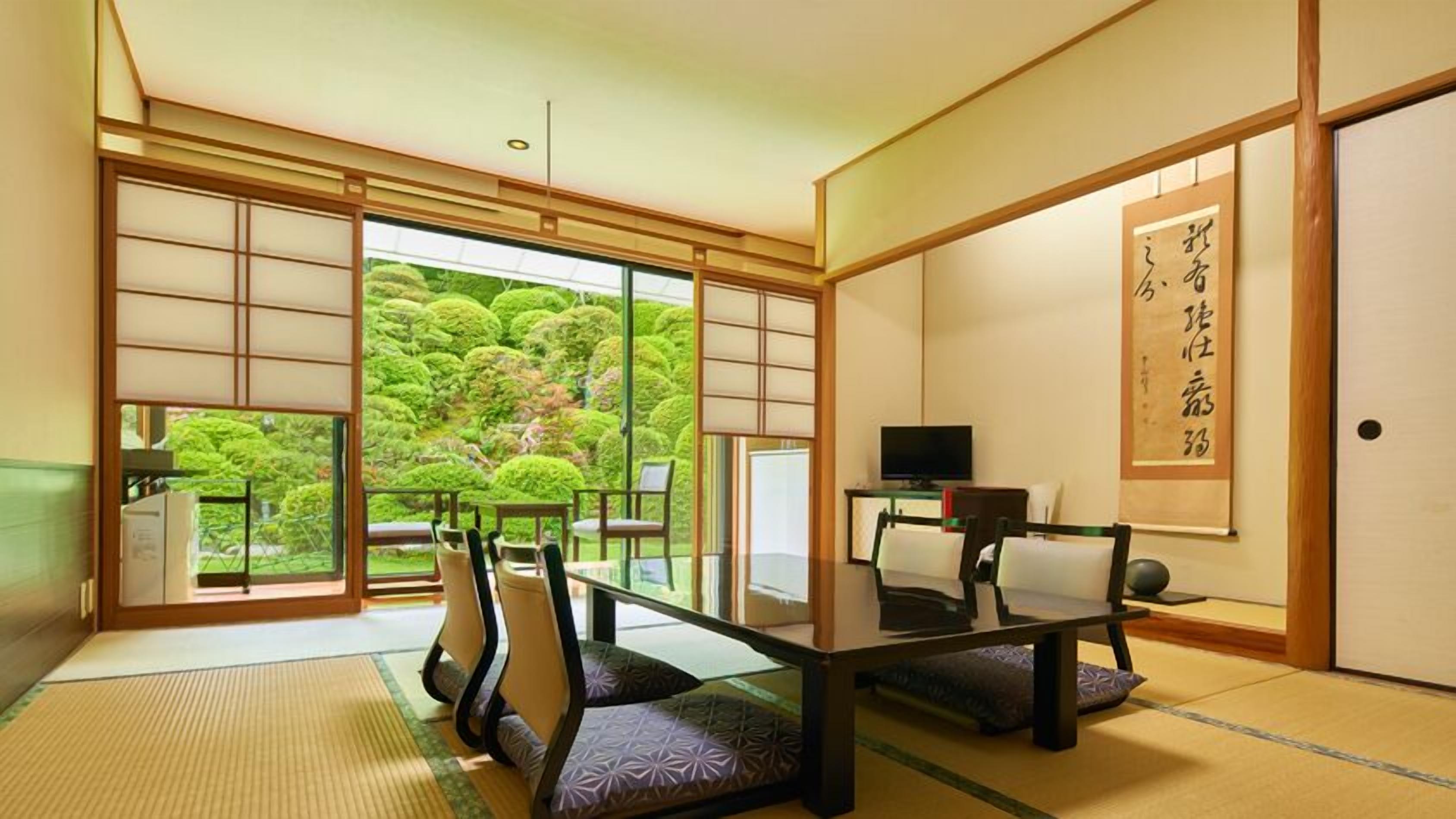 Habitació japonesa tradicional amb fons de tatami, una taula baixa, cadires, televisió i una finestra gran amb vista a un jardí exuberant.