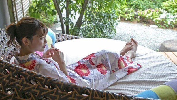 Une personne allongée sur une chaise ronde en osier tressé, recouverte d’une couverture à motifs, à l’extérieur, avec de la verdure en arrière-plan.