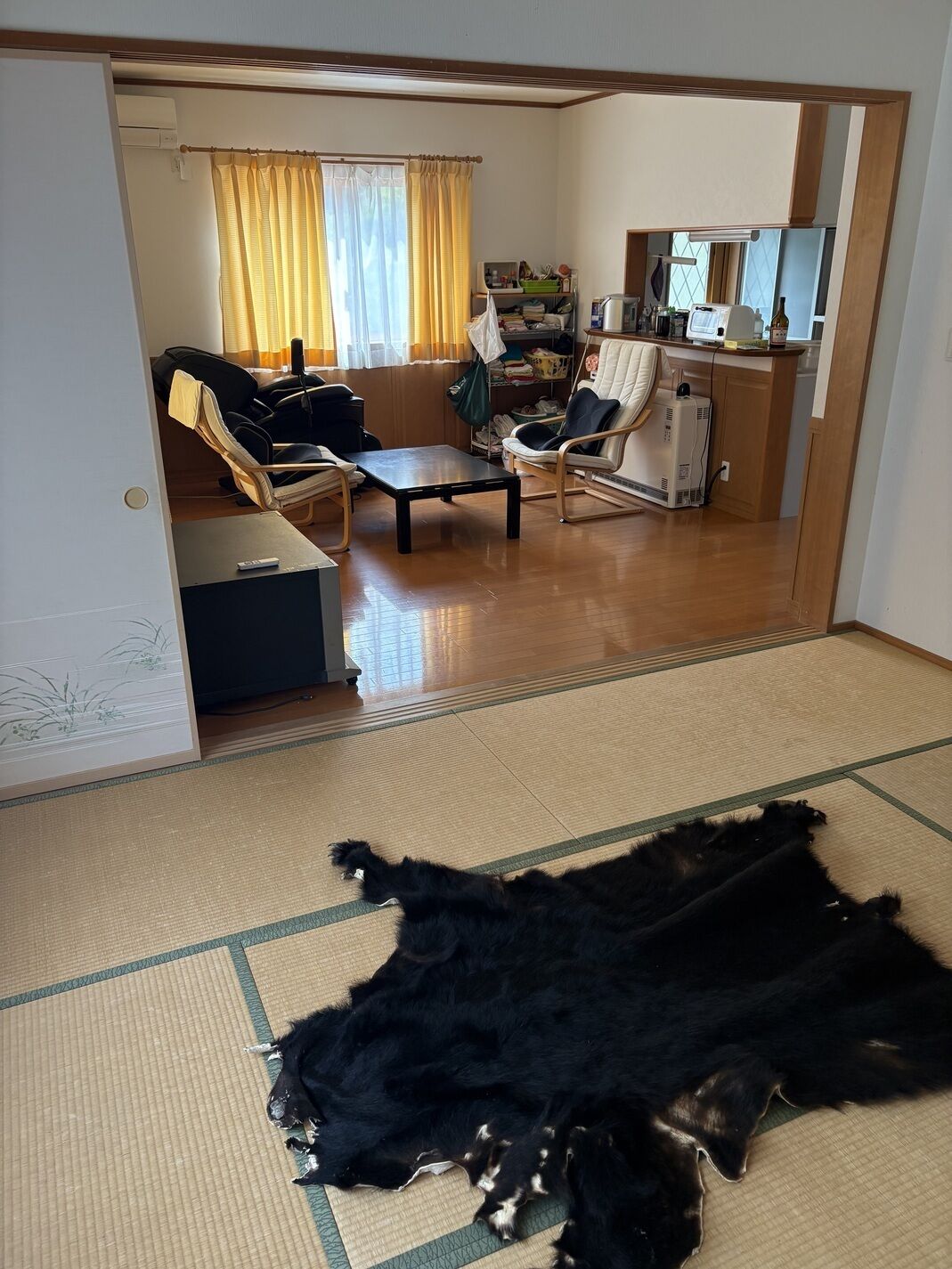 Ein Zimmer mit Tatami-Matte, einem schwarzen Hund, einem Stuhl, einem Tisch und einem Regal mit verschiedenen Gegenständen.