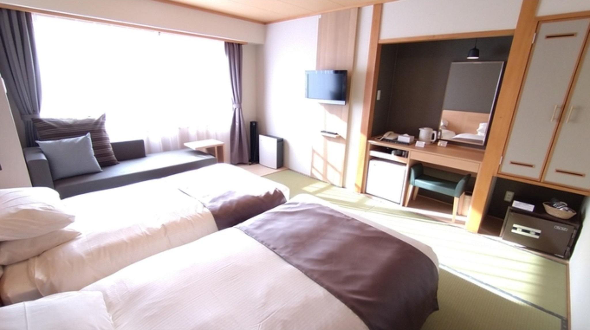 A hotel room with two beds, a desk, a TV, and a kitchenette.