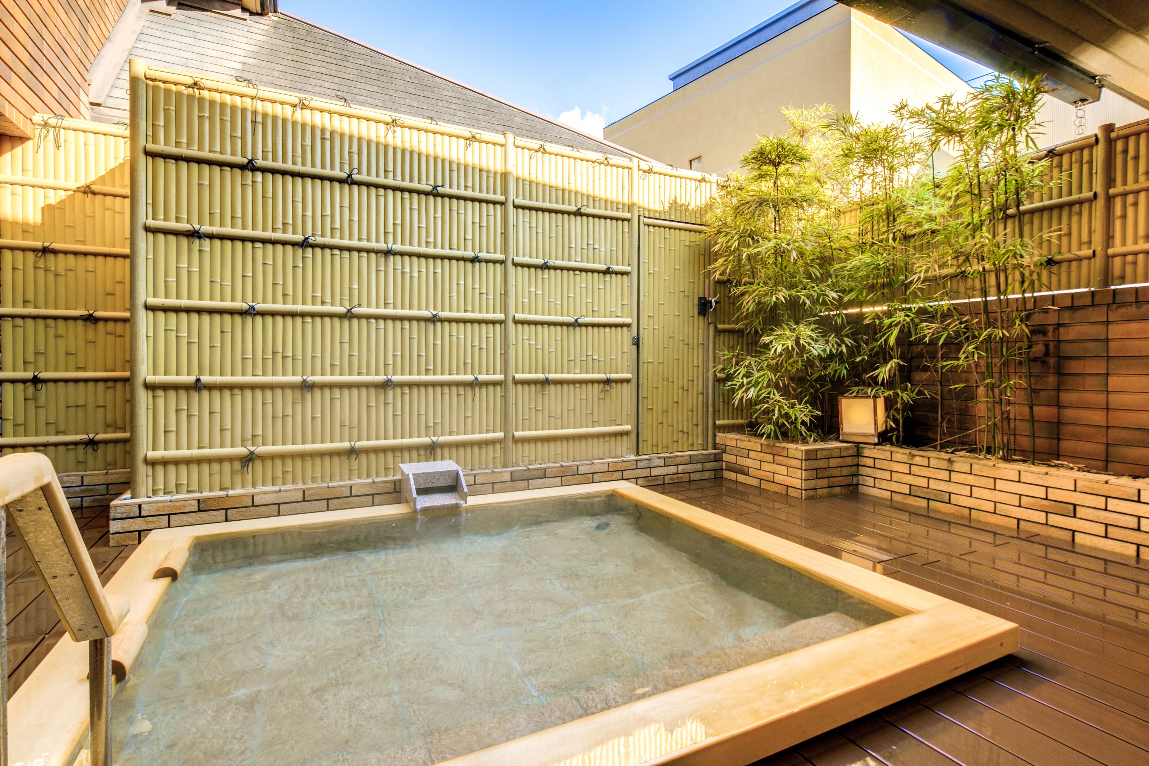A traditional Japanese onsen with a wooden deck, bamboo wall, and a brick wall with a bamboo plant.