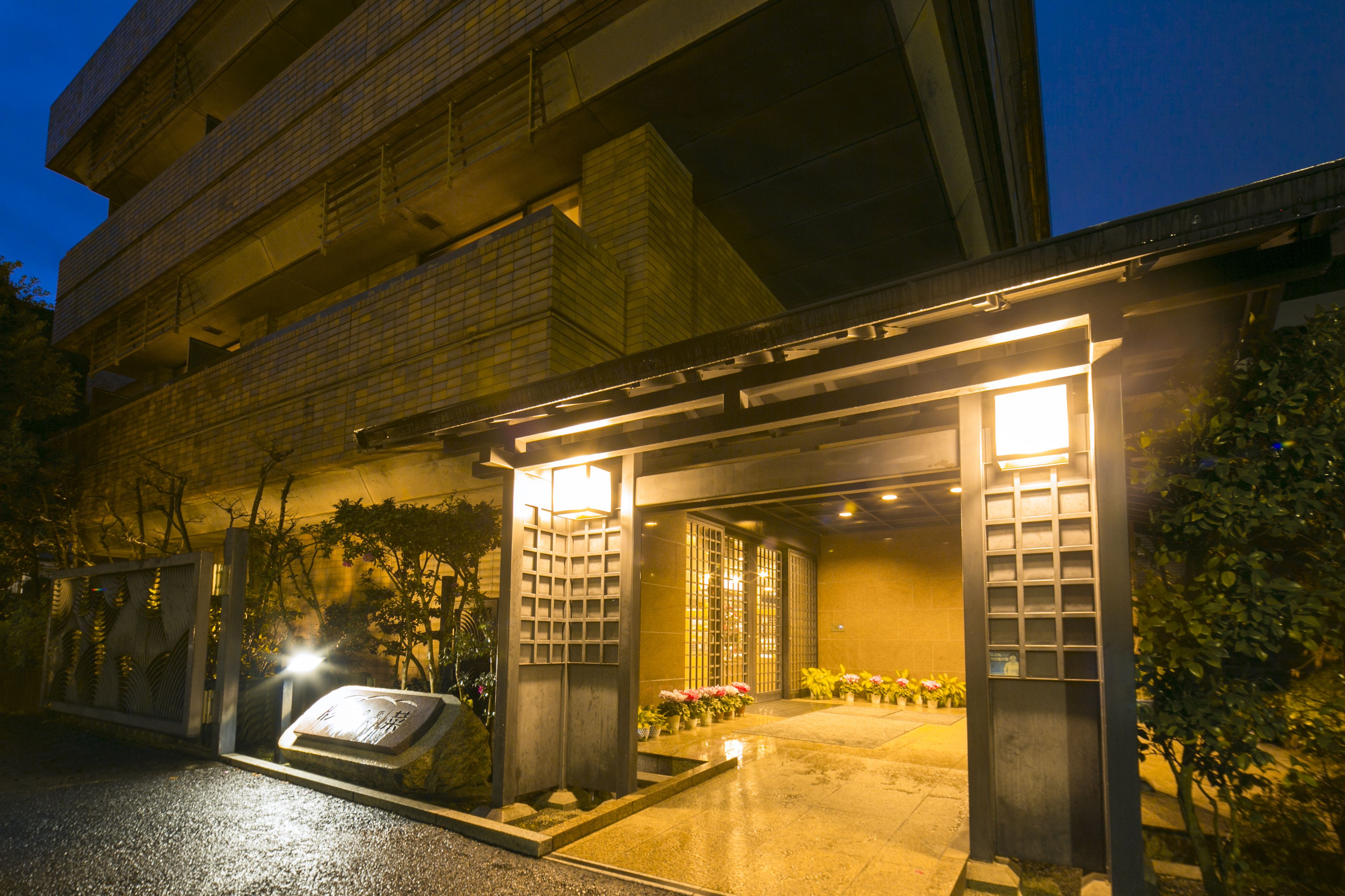 Entrada de hotel con fachada de azulejos, un área de acceso cubierta y un pequeño jardín con flores.