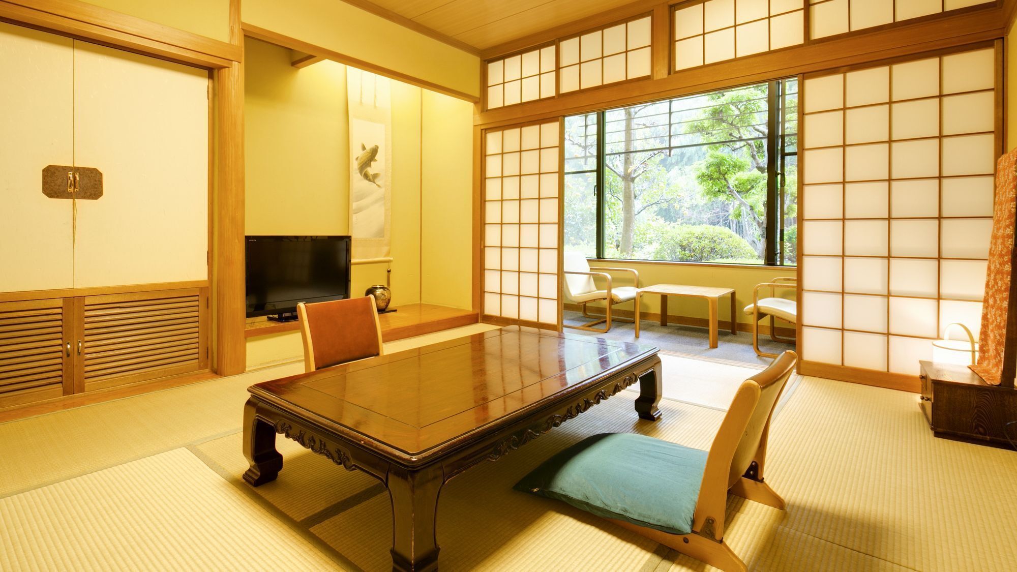 A traditional Japanese room with tatami flooring, a low table, chairs, a television, and a sliding door with a view of greenery.