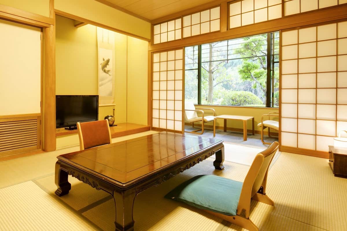 Una habitación tradicional japonesa con suelo de tatami, una mesa baja, sillas, un televisor y una puerta corrediza con vistas a la vegetación.
