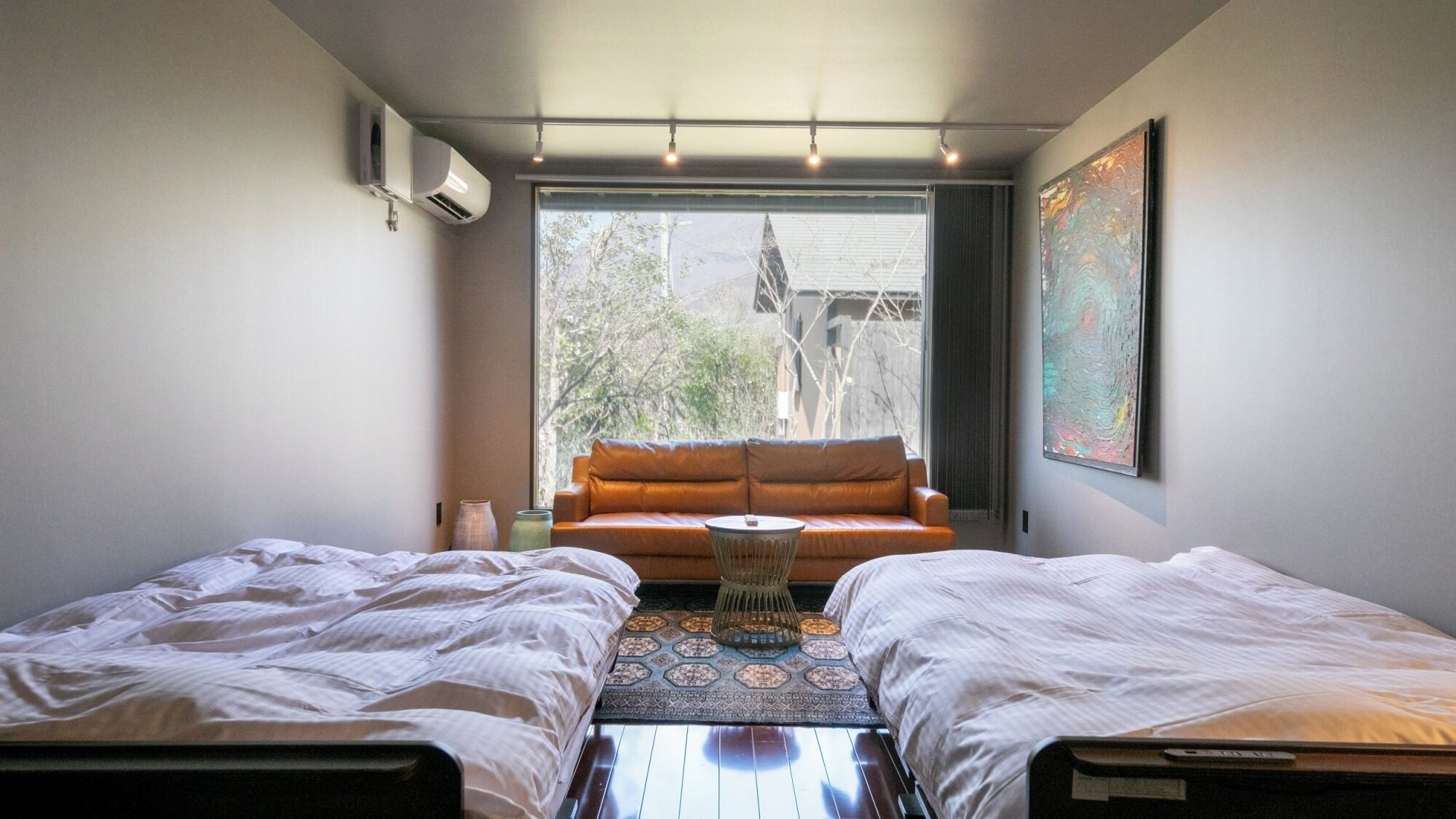 Ein Zimmer mit zwei Betten, einem Schlafsofa, einem kleinen Tisch und einem großen Fenster mit Blick auf Gebäude und Bäume.