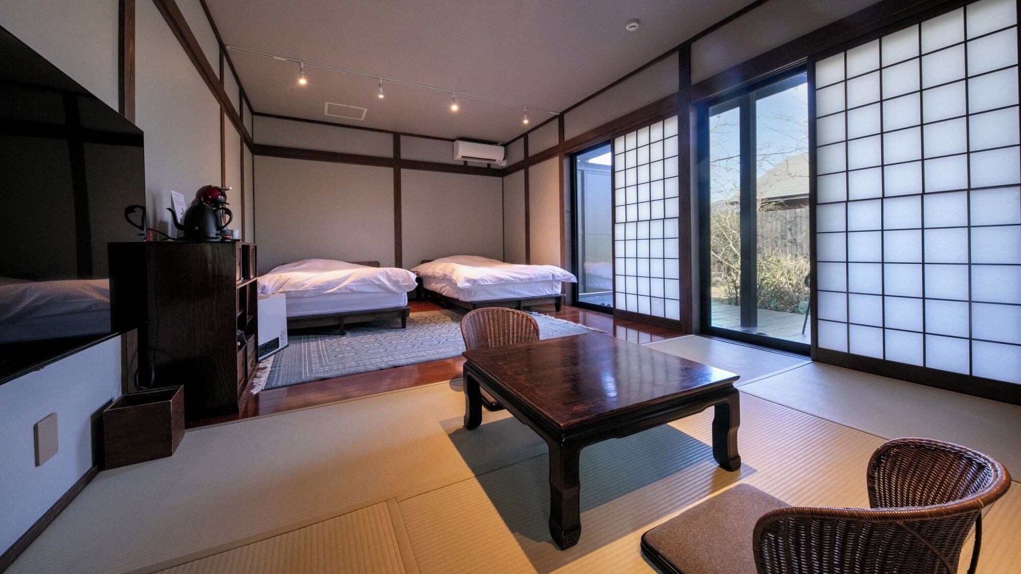 Ein traditioneller japanischer Raum mit Tatami-Boden, einem niedrigen Holztisch, zwei Betten und einer Schiebeglastür, die zu einem Garten führt.