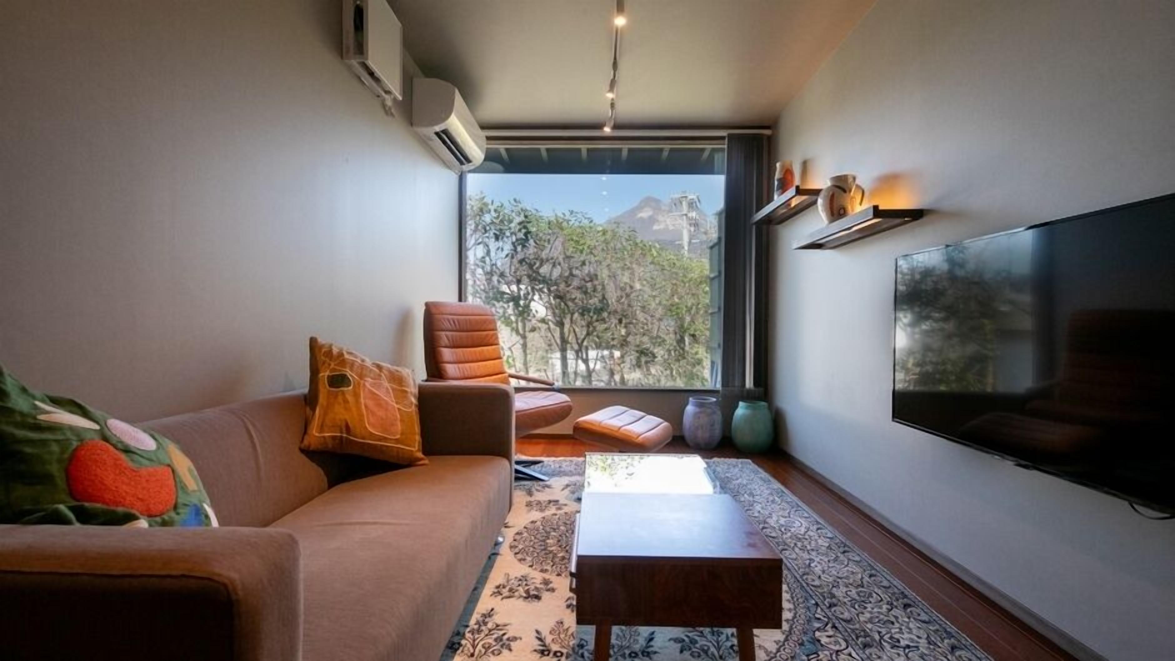 A modern living room with a sofa, a chair, a small table, and a large TV. There is a window with a view of trees and a mountain.