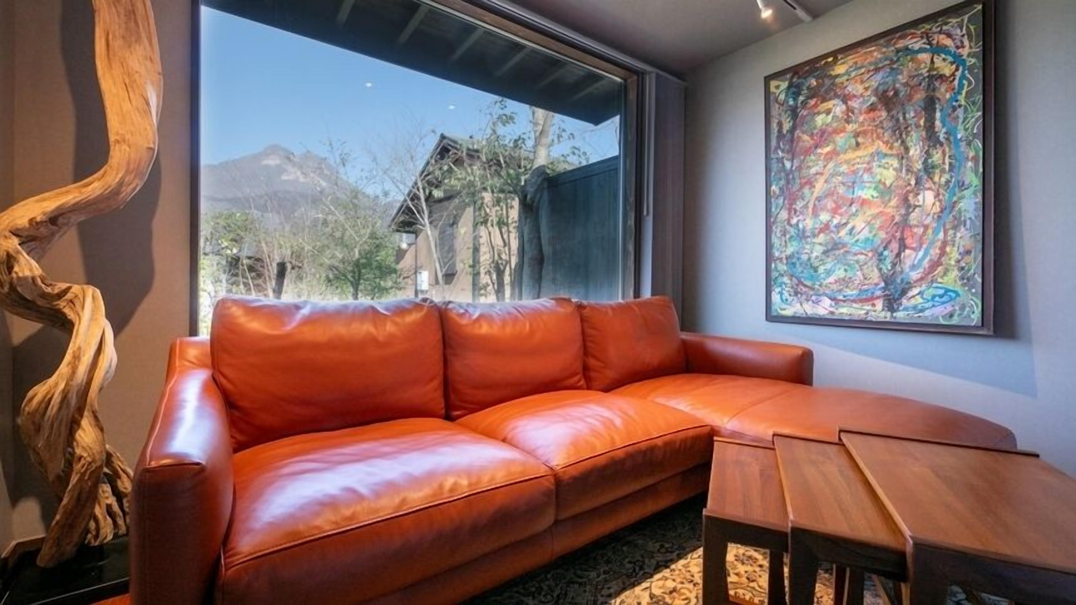 A leather sofa, a wooden table, and a large abstract painting in a room with a view of a mountain and trees.