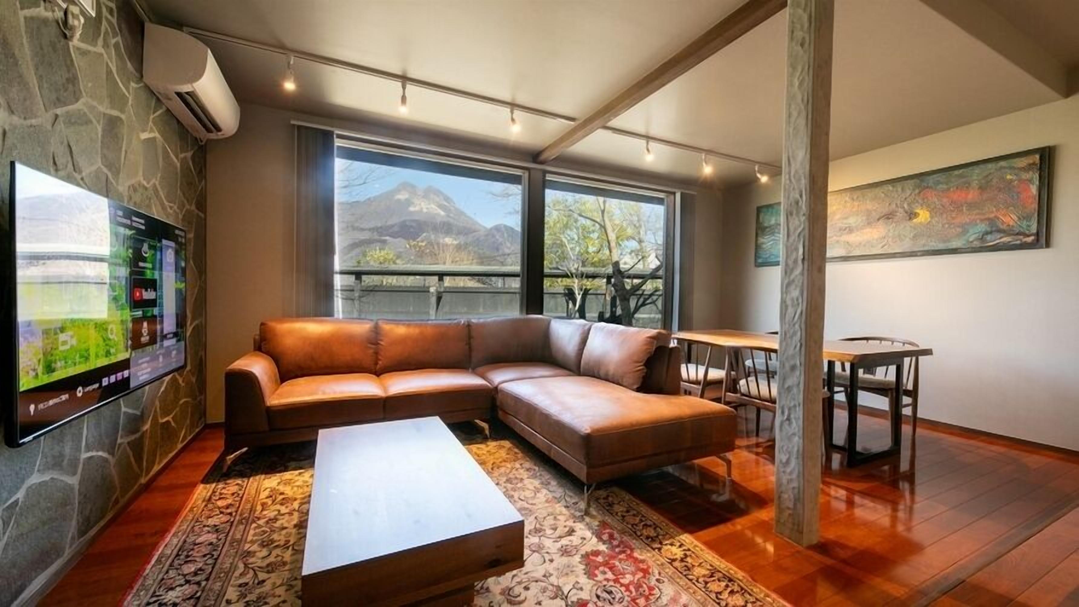 A modern living room with a leather sofa, a coffee table, a TV mounted on a stone wall, and large windows offering a mountain view.