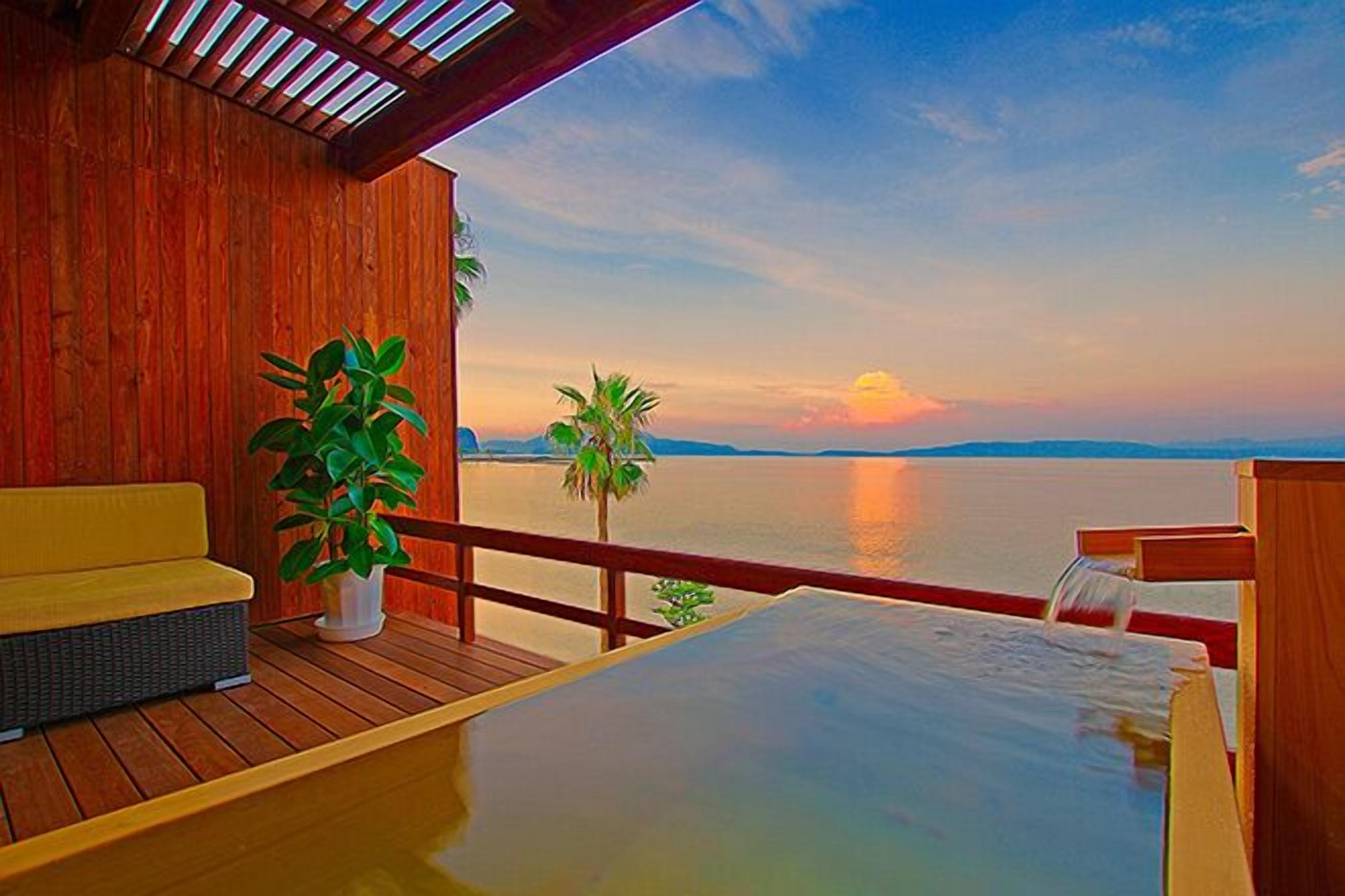 Une terrasse en bois avec un canapé, une plante en pot et un jacuzzi, le tout offrant une vue sur une étendue d’eau au coucher du soleil.